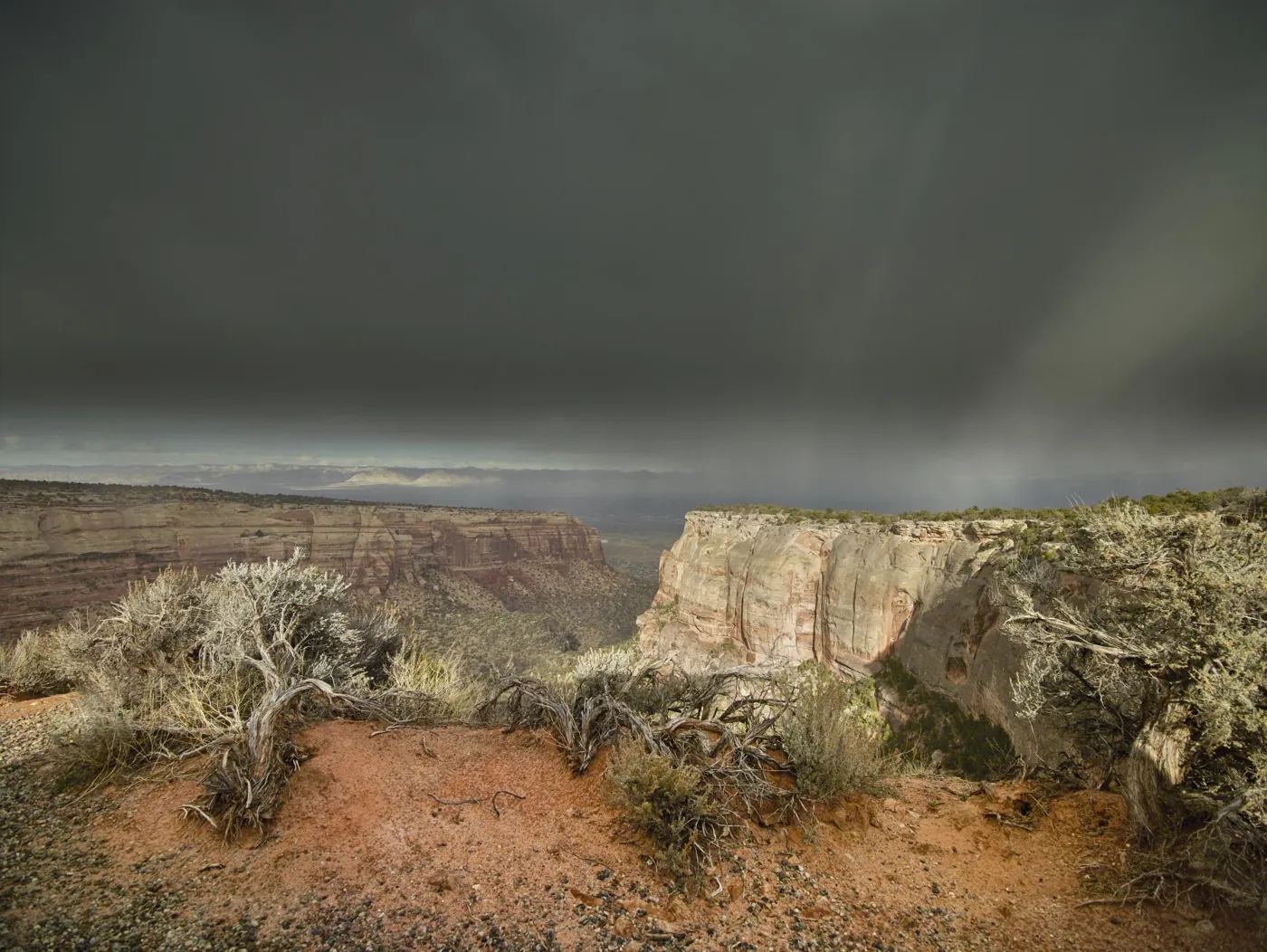 Storm clouds above sandstone canyon walls and a juniper-lined rim at Colorado National Monument
